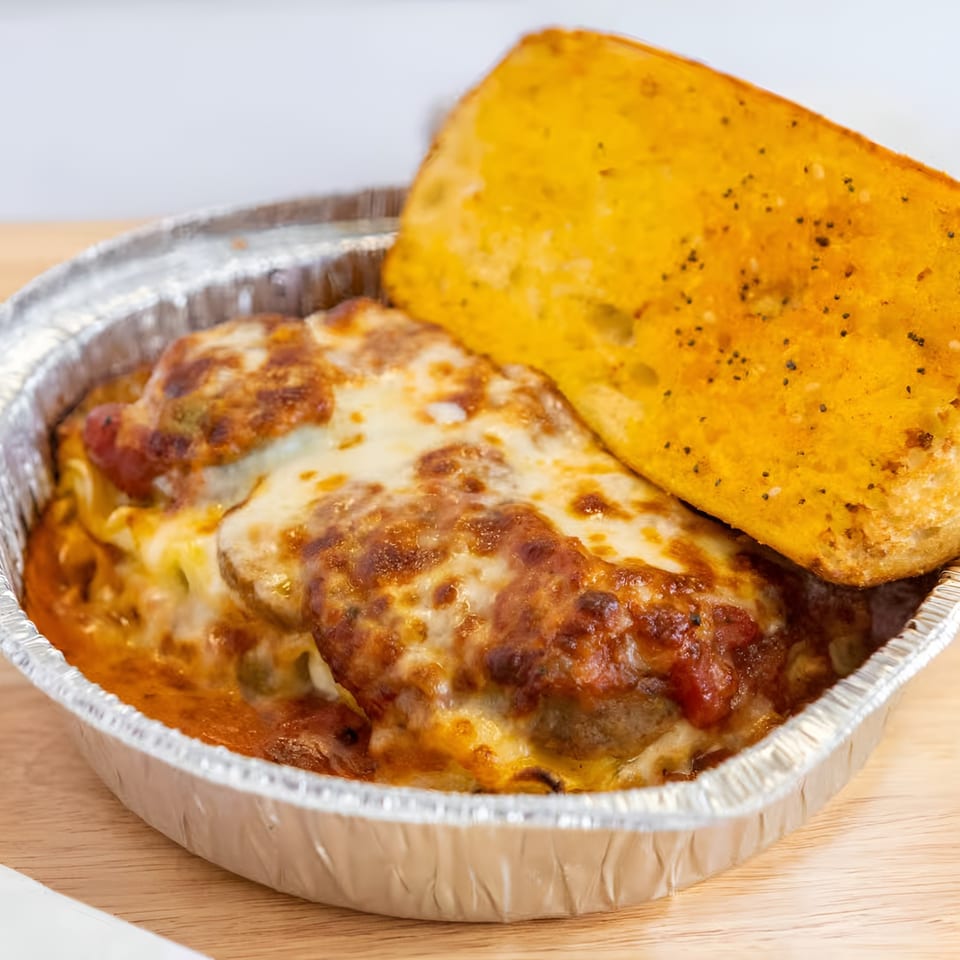 Baked pasta with melted cheese and tomato sauce in a foil container, with a side of garlic bread.