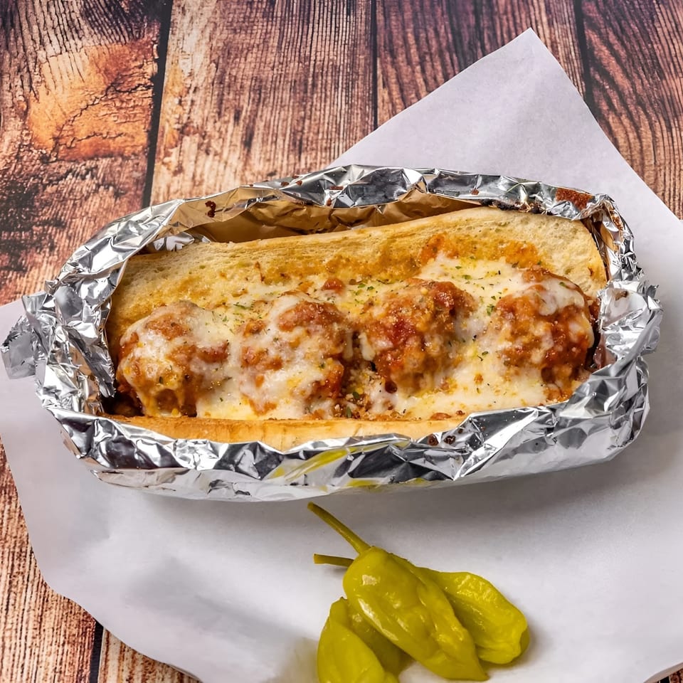 Meatball sub with melted cheese and sauce in foil, served with green peppers on white paper.