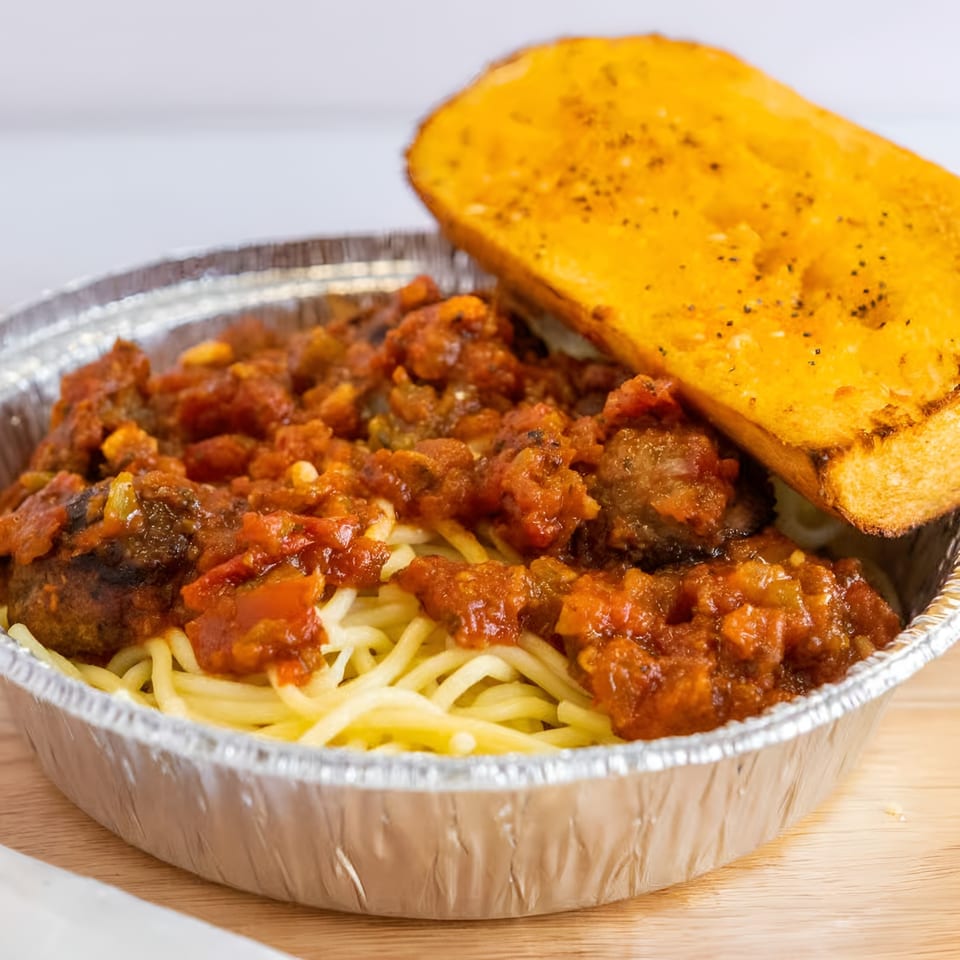 Spaghetti with meatballs and tomato sauce in a foil container, served with a slice of garlic bread.